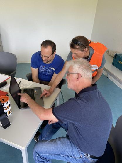 Das Bild zeigt drei Personen, die um einen Tisch sitzen und gemeinsam an einem Laptop arbeiten. Der Mann im Vordergrund trägt ein dunkelblaues Poloshirt und zeigt auf den Bildschirm des Laptops. Er hat graues Haar und trägt eine Brille. Der Mann links trägt ein blaues T-Shirt mit einem weißen Muster und hat kurze, dunkle Haare. Der Mann in der Mitte trägt eine orangefarbene Weste mit dem Aufdruck "RSKN" und hat eine Sonnenbrille auf dem Kopf. Er beugt sich über den Laptop und scheint auf den Bildschirm zu schauen. Auf dem Tisch befinden sich ein Laptop, ein Notizbuch, ein Stift und eine Spendendose. Der Raum hat eine einfache, funktionale Einrichtung mit grauen Stühlen und einem hellen Tisch.