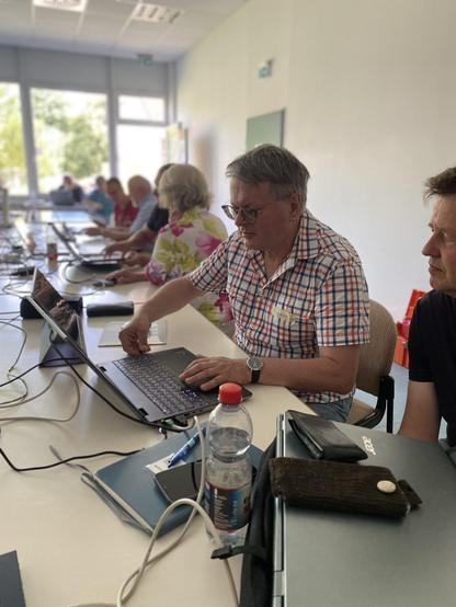 Das Bild zeigt eine Gruppe von Menschen, die an einem langen Tisch sitzen und auf ihren Laptops arbeiten. Im Vordergrund ist ein Mann in einem karierten Hemd zu sehen, der auf seinem Laptop tippt. Neben ihm steht eine Wasserflasche und ein Notizbuch. Auf dem Tisch liegen verschiedene Gegenstände, darunter ein schwarzer Geldbeutel und ein schwarzes Lederetui. Im Hintergrund sitzen weitere Personen, die ebenfalls auf ihren Laptops arbeiten. Die Umgebung wirkt hell und freundlich, mit großen Fenstern, die viel Licht hereinlassen.