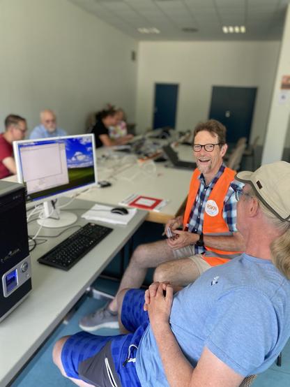 Das Bild zeigt eine Gruppe von Menschen in einem Raum, der wie ein Computerlernraum oder ein Technikworkshop aussieht. Im Vordergrund sitzen zwei Männer an einem Tisch. Der Mann auf der linken Seite trägt eine orangefarbene Weste mit einem Aufkleber, der "RSKN" zeigt, und eine Brille. Er lächelt und scheint mit dem Mann auf der rechten Seite zu sprechen, der eine beige Kappe und ein blaues T-Shirt trägt. Der Mann auf der rechten Seite hat die Arme verschränkt und blickt den anderen Mann an. Auf dem Tisch vor ihnen stehen ein Computer mit einem Monitor, einer Tastatur und einem Mauspad. Im Hintergrund sitzen weitere Personen an den Tischen, die ebenfalls an Computern arbeiten. Die Wände sind weiß, und es gibt zwei Türen im Hintergrund. Die Atmosphäre wirkt freundlich und informell.
