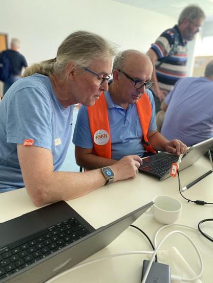 Zwei Männer sitzen an einem Tisch und arbeiten an ihren Laptops. Der Mann auf der linken Seite trägt ein hellblaues T-Shirt und hat langes, graues Haar, das zu einem Pferdeschwanz gebunden ist. Er trägt eine Armbanduhr und hat eine Namensschilder auf seinem Shirt. Der Mann auf der rechten Seite trägt ein blaues Poloshirt und eine orangefarbene Weste mit einem "RSKN" Logo. Er hat kurzes, graues Haar und trägt eine Armbanduhr. Beide Männer konzentrieren sich auf ihre Laptops, der Mann auf der rechten Seite tippt auf der Tastatur. Auf dem Tisch liegen mehrere Laptops, ein weißer Becher und Kabel. Im Hintergrund sind weitere Personen zu sehen, die ebenfalls an der Aktivität teilnehmen.