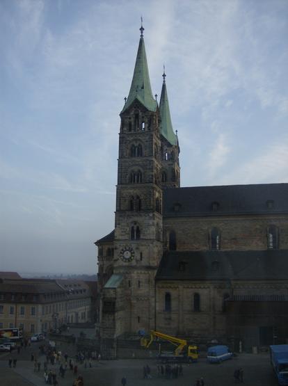 The two spires of Bamberger Dom seen almost directly from the side, with one barely showing from behind the other. The surrounding square is dark and the clouds overhead are lightly illuminated by the setting sun.