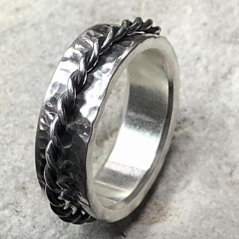A silver ring featuring a hammered texture, with a twisted metal detail running along its edge. The background is neutral and textured.
