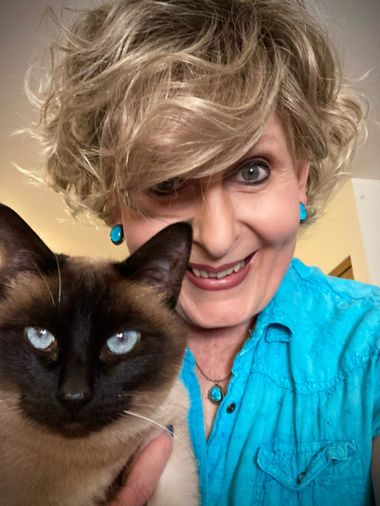An older woman with blonde hair is wearing turquoise jewelry and a top that’s the same color. Beside her, on the left, is a Siamese cat with blue eyes who is staring at the camera.