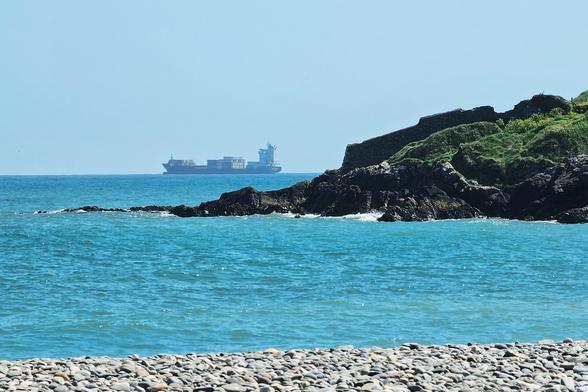 A wide shot of the Bray coastline on a clear day, featuring the shimmering blue sea in the foreground and a rocky outcrop on the right. In the distance, a large cargo ship laden with containers sails across the horizon under a pale blue sky. A pebble beach is visible at the very bottom of the frame.