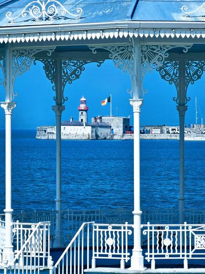 A bright blue seascape with a lighthouse and harbour visible through the white, ornate pillars and railings of a Victorian-style bandstand or pavilion. The water is a deep blue, and the sky is clear. The red and white lighthouse stands prominently on a stone pier, with the Irish flag flying nearby.