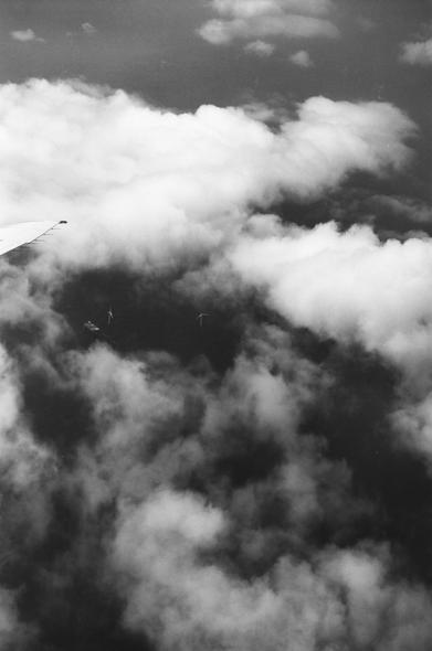 A black and white analog photo taken from an airplane window. The image shows a small part of the plane wing, clouds, and the sea with windmills and a ship.