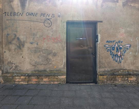 A sunlit, weathered concrete wall with a dark grey metal door in the centre, stretching from a brick base to the top of the frame. The wall is covered in various graffiti: to the left of the door, "1 LEBEN OHNE PENG" is written in black, with a simple stick figure drawing below it. Further left and below that, faint red scribbles are visible. To the right of the door, a blue, pink, and white graffiti tag resembles a winged, skull-like creature with "KAUF & P" written above it. The door itself has a horizontal letterbox slot and a handle. Below the wall, a dark grey paved footpath extends across the foreground. Sunlight creates a flare effect in the top right corner of the image.