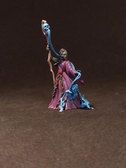 Das Bild zeigt eine detaillierte Miniaturfigur, die einen magischen Wesen darstellt. Die Figur steht auf einem dunklen Hintergrund und trägt ein langes, purpurfarbenes Gewand mit einem roten Gürtel. Ihr Gesicht ist grün, und sie hat lange, dunkle Haare, die in Zöpfen geflochten sind. In ihrer rechten Hand hält sie einen langen, gebogenen Zauberstab, der mit einem blauen Schädel verziert ist. Die linke Hand der Figur ist erhoben, und aus ihrem Arm wächst ein blauer, geschmeidiger Arm, der einen kleineren, blauen Geist oder Wesen umarmt. Die Figur strahlt eine mystische und mächtige Aura aus, die durch die Farben und die detaillierte Malerei unterstrichen wird.