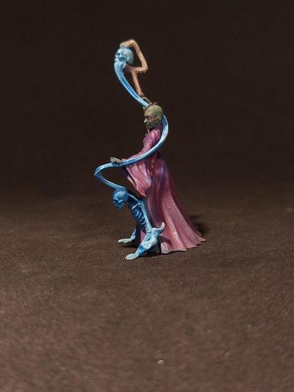 Das Bild zeigt eine detaillierte Miniaturfigur, die einen magischen Wesen darstellt. Die Figur steht auf einem dunklen Hintergrund und trägt ein langes, purpurfarbenes Gewand mit einem roten Gürtel. Ihr Gesicht ist grün, und sie hat lange, dunkle Haare, die in Zöpfen geflochten sind. In ihrer rechten Hand hält sie einen langen, gebogenen Zauberstab, der mit einem blauen Schädel verziert ist. Die linke Hand der Figur ist erhoben, und aus ihrem Arm wächst ein blauer, geschmeidiger Arm, der einen kleineren, blauen Geist oder Wesen umarmt. Die Figur strahlt eine mystische und mächtige Aura aus, die durch die Farben und die detaillierte Malerei unterstrichen wird.