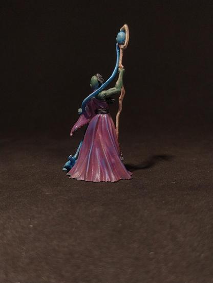 Das Bild zeigt eine detaillierte Miniaturfigur, die einen magischen Wesen darstellt. Die Figur steht auf einem dunklen Hintergrund und trägt ein langes, purpurfarbenes Gewand mit einem roten Gürtel. Ihr Gesicht ist grün, und sie hat lange, dunkle Haare, die in Zöpfen geflochten sind. In ihrer rechten Hand hält sie einen langen, gebogenen Zauberstab, der mit einem blauen Schädel verziert ist. Die linke Hand der Figur ist erhoben, und aus ihrem Arm wächst ein blauer, geschmeidiger Arm, der einen kleineren, blauen Geist oder Wesen umarmt. Die Figur strahlt eine mystische und mächtige Aura aus, die durch die Farben und die detaillierte Malerei unterstrichen wird.