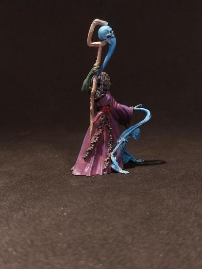 Das Bild zeigt eine detaillierte Miniaturfigur, die einen magischen Wesen darstellt. Die Figur steht auf einem dunklen Hintergrund und trägt ein langes, purpurfarbenes Gewand mit einem roten Gürtel. Ihr Gesicht ist grün, und sie hat lange, dunkle Haare, die in Zöpfen geflochten sind. In ihrer rechten Hand hält sie einen langen, gebogenen Zauberstab, der mit einem blauen Schädel verziert ist. Die linke Hand der Figur ist erhoben, und aus ihrem Arm wächst ein blauer, geschmeidiger Arm, der einen kleineren, blauen Geist oder Wesen umarmt. Die Figur strahlt eine mystische und mächtige Aura aus, die durch die Farben und die detaillierte Malerei unterstrichen wird.