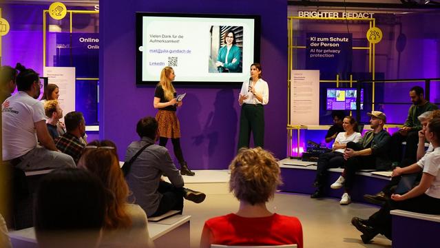 Nora Zupan und Julia Gundlach bei ihrem Vortrag beim Civic Coding x Mission KI Forum. Sie stehen vor dem Plenum und einem Bildschirm, der Informationen zu Julia Gundlach zeigt. Im Publikum sitzen verschiedene Personen, die interessiert zuhören.