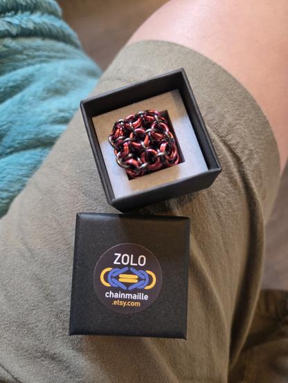 A pink and black chainmaille fidget cube in a grey 3D printed insert in a black jewellery box. The lid of the box is shown beneath it, with a sticker on it. The sticker is black and shows the words ZOLO and chainmaille in white bracketing a stylised Byzantine weave in yellow and blue, beneath the word chainmaille is .etsy.com in yellow