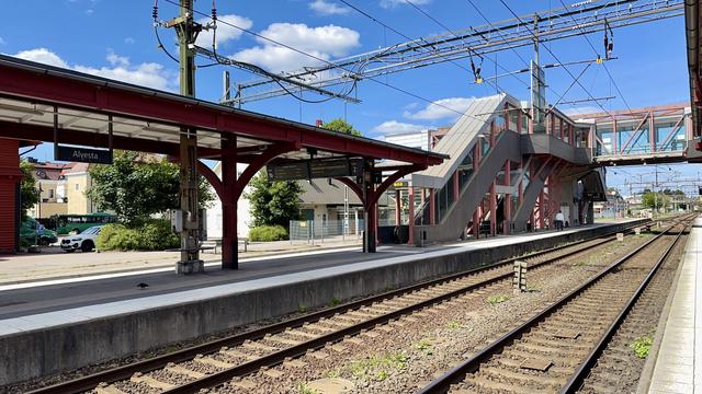 A train station platform marked "Alvesta," featuring a red roof and waiting area. A staircase leads to an overhead bridge, with tracks visible in the foreground and buildings in the background under a blue sky with clouds.