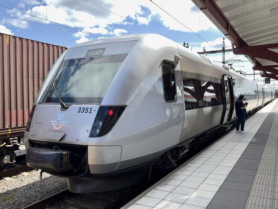 A silver train, likely a regional or high-speed service, is parked at a station. The train has a prominent front design and the number "3351" displayed. A person stands on the platform, checking their phone, with shipping containers visible.