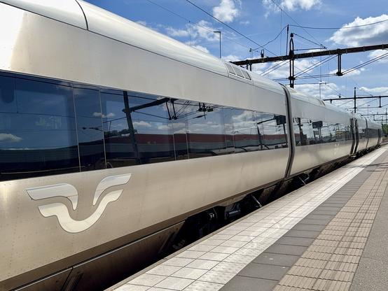 A sleek, modern train is parked at a station platform under a blue sky with clouds. The train features a metallic exterior with a distinct logo visible. Electric overhead lines and other train infrastructure are present in the background.