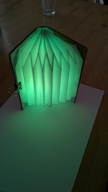 a half opened DIY book lamp. Light-up green tinting the table and a white sheet of paper in green light. The book lamp stands upright spreading its bellows to the camera. Camera is at about 45 degree angle.