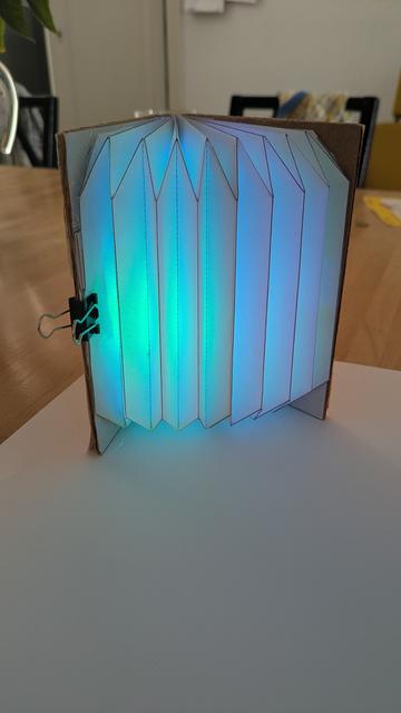 a half opened DIY book lamp. Light-up teal and green, tinting the table and a white sheet of paper in blueish green light. The book lamp stands upright spreading its bellows to the camera. Camera is at about 45 degree angle.