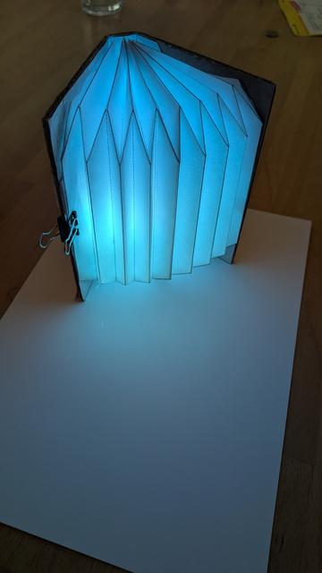 a half opened DIY book lamp. Light-up blue, tinting the table and a white sheet of paper in blue light. The book lamp stands upright spreading its bellows to the camera. Camera is at about 45 degree angle.