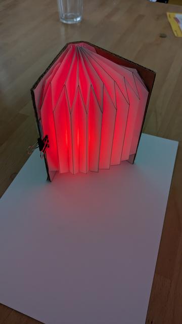 a half opened DIY book lamp. Light-up red tinting the table and a white sheet of paper in red light. The book lamp stands upright spreading its bellows to the camera. Camera is at about 45 degree angle.