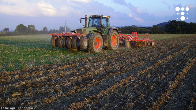 Traktor mit Anbaugerät bearbeitet Ackerland unter blauem Himmel mit Wolken, im Hintergrund Bäume und Hügel