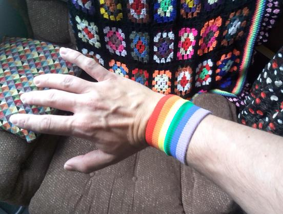 A photo of my outstretched hand with a knitted rainbow wristband.  The background is a brown fabric armchair with a multicoloured pillow and a crocheted multicolour blanket.