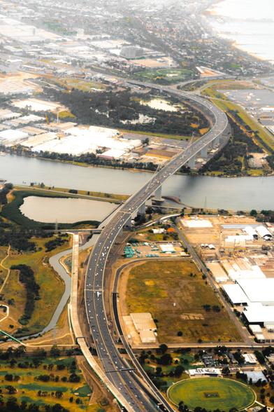 Aerial view of a highway bridge spanning a wide river, connecting two industrial areas. The bridge curves slightly, with vehicles visible along its length. Surrounding land includes green patches, buildings, and smaller roads. The distant background shows more urban development and water bodies.