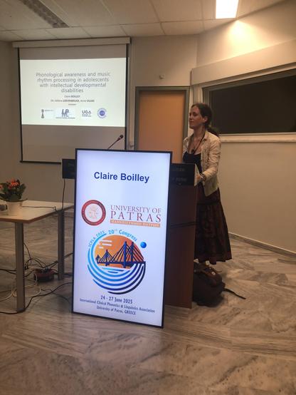Me, a white woman in her thirties, with long hair, dressed in a long black and orange skirt and a white shirt, standing in front of a desk with a microphone and a screen showing my name above the logo of the university of Patras. Behind me, the first slide of my presentation is being projected, showing the title: "phonological awareness and music rhythm processing in adolescents with intellectuel developmental disabilities".