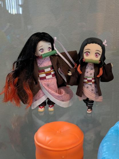 Two small dolls of Nezuko Kamado, a character from the anime Kimetsu no Yaiba/Demon Slayer. The doll on the left is an Azone International DOLPokke with brushable hair and acrylic "glass" eyes while the other is a Nendoroid Doll with plastic hair and a plastic faceplate. Both are wearing pink kimono.

There is plastic Barbie furniture in the foreground because I was in a hurry and didn't take the time to stage this photo properly.