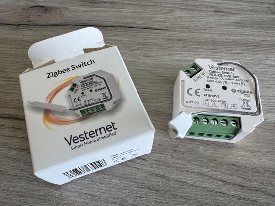 Image features a Zigbee switch from Vesternet, displayed alongside its packaging. The switch is white with green terminals, labeled with technical specifications.