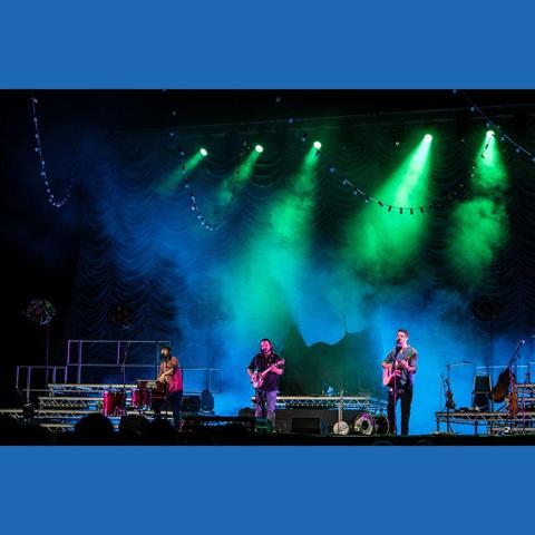 A photo of your pals and mine, The Longest Johns, on a stage lit up with green spotlights and blue smoke, while Robbie plays the Harmonium, Andy's playing banjo and singing, and JD plays the guitar also whilst singing.