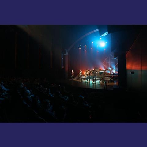 A distant photo of Home Free on a stage, taken from the position of the audience. It shows the band lined up across the stage, in the middle of a song. The backdrop of the stage has a red/orange glow, and there's a cold blue spotlight above them shining out towards the audience.