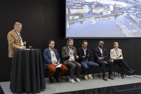 Das Foto zeigt eine Podiumsdiskussion mit sechs Personen auf einer Bühne. Links steht ein Mann mit Mikrofon hinter einem Stehtisch, er trägt ein beigefarbenes Sakko. Daneben sitzen fünf weitere Personen auf Stühlen. In der Mitte spricht ein Mann mit grauem Sakko und lockigen Haaren ins Mikrofon. Es ist der Forschungsdirektor der Cyberagentur, Prof. Dr. Christian Hummert. Die anderen hören aufmerksam zu. Im Hintergrund ist ein großes Projektionsbild mit einer Luftaufnahme der Stadt Helsinki und einem Hafen mit Segelbooten zu sehen. Die Teilnehmer tragen Namensschilder und blaue Konferenzbändchen. Die Stimmung wirkt konzentriert, aber freundlich – typisch für eine internationale Konferenz im Innovationsumfeld. Foto: © Susanna Lehto