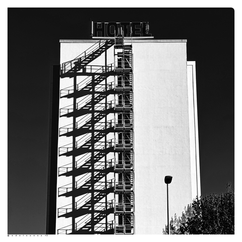 Schwarzweißfotografie: Seitenansicht von einem hellen Hochhaus mit Großbuchstaben auf dem Flachdach „HOTEL“, die mittig angebrachte Feuerleiter erstreckt sich über alle Etagen, links davon auf der glatten Fassade, der entsprechende verzerrte dunkle Schatten von Treppen und Zwischenetagen