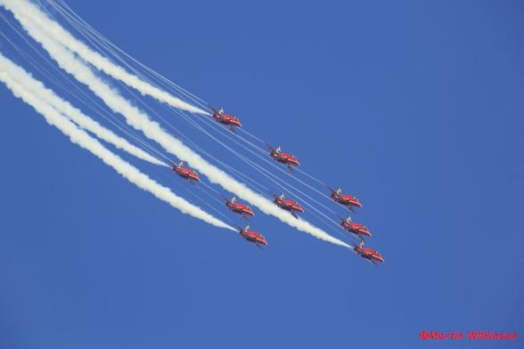 The Royal Air Force Red Arrows display team fly in formation through a clear blue sky at an air display.