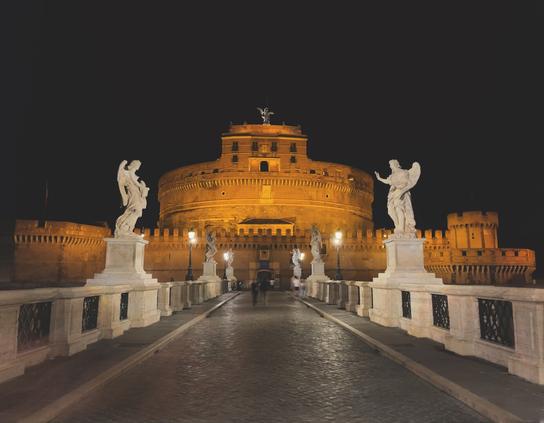 Night view of Castel Sant'Angelo in Rome, illuminated by warm lights. The foreground features statues of angels on either side of a stone bridge, with a cobblestone walkway and soft streetlights.