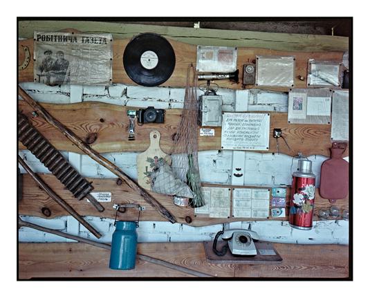 A wooden wall covered with vintage Soviet-era objects: an old rotary phone, a vinyl record, a gas mask filter, a film camera, thermos, newspaper clippings, and handwritten signs. Captured on expired Kodak Portra 160 (2022) using a Fuji GA645w medium format camera.