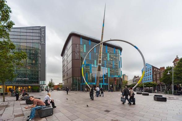 View of an urban plaza filled with passersby. At the back is a library, a tall building with rounded corners and a glass facade with vertical coloured panels. In front of the library is a large art installation of a pointed spire and partially buried hoop.