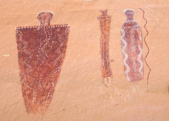 Four red painted figures with white embellishments on a tan cliff wall. Two forms are human like, one is animal like, and one is a long vertical zigzag red and white snake.