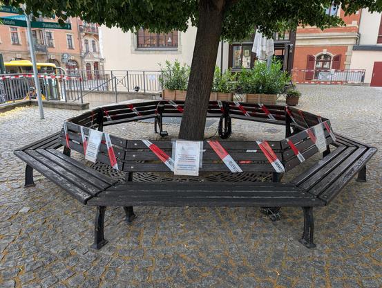 Das Foto zeigt eine Bank, die einen Baum unter seiner Baumkrone umringt. Die Bank ist mit Absperrband umwickelt, welches die Aufschrift Feuerwerk-Abbrennplatz-Sperrzone trägt. Außerdem gibt es Zettel mit der Aufschrift "Am Samstag 28.06.2025 ist der Platz wegen Open Air Veranstaltung ab 12:00 Uhr gesperrt! Etwaige angeschlossene Fahrräder werden kostenpflichtig entfernt."