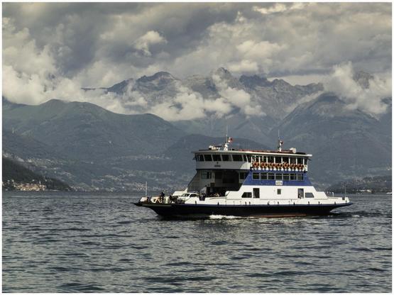 A ferry named "Aida" navigates a lake surrounded by green mountains and cloudy skies, with several passengers visible on board.