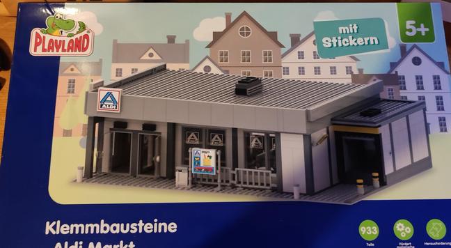 Picture of a packaging for a Clip brick construction set for kids similar to Lego. It's called Playland showing a display like shop of an Aldi Nord Store.

The store looks like the newest modernized Aldi stores you can find everywhere in the north of Germany.

On the packages a blue frame reaches from the left upper corner with, down into a round 90° curve to the bottom right, in white letters on this blue frame it reads, in German, Klemmbausteine - Aldi-Markt.
On the right lower corner 3 green circles with white text read 933 Teile, fördert motorische Fertigkeiten & Herausforderung.

The Modell itself is portrait with the front, the right side and the roof visible above the blue frame, simple depictions of, middle sized, city houses are behind it and the Modell is placed on a white grey ground, contrasting it's mid and dark grey tones and some blue elements.
The upper right corner has a green flag with a white 5+ for the age requirement and next to it on the left an also green rectangle with slightly tilted sides and tilted text mit Stickern.