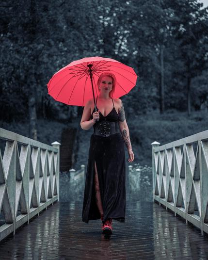 A woman with red hair walks in the rain holding a bright red umbrella. She is wearing a black dress with a thigh-high slit and red boots. The bridge has white railings with a crisscross pattern and is surrounded by blurred trees in the background.