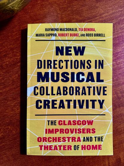 A yellow covered paperback on a wooden table. The book is New Directions in Musical Collaborative Creativity - The Glasgow Improvisers Orchestra and the Theater of Home, by Raymond MacDonald, Tia DeNora, Maria Sappho, Robert Burke and Ross Birrell.