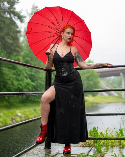 Red-haired woman stands on a wet platform holding a large red umbrella. Wears a black dress with a high slit and red boots. Background features greenery and a river. Overcast sky suggests rain, with visible raindrops. A bridge is visible in the distance.
