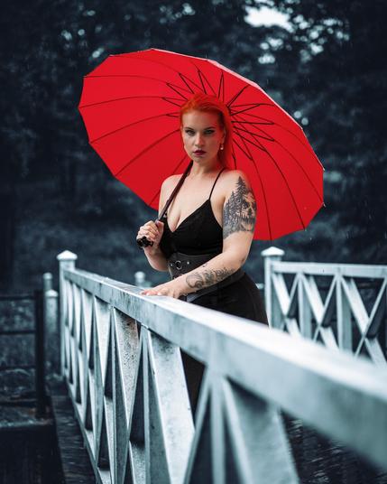 Woman with red hair and tattoos holds a vibrant red umbrella while standing on a metal bridge. She wears a black sleeveless dress with thin straps. The background is a dark, rainy forest with blurred tree details. The bridge has a repeating triangular pattern along its railing.