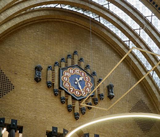 Vue en contre-plongée d'un grand mur intérieur en brique jaune dont le haut en demi-cercle est rattaché à une verrière. Une grande horloge est installée contre ce mur. Elle est posée sur des colonnes de briques jaunes parallèles formant un losange et se terminant à chaque extrémité par un élément rond noir. Deux autres losanges de part et d’autre viennent équilibrer le tout. On aperçoit aussi un morceau d'éclairage sous forme d'un cercle lumineux suspendu en hauteur.