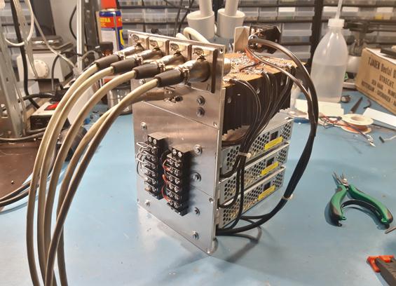 An electronic device on a working bench with component drawers, some tools and a microscope in the background. The device is metal-coloured, an electronics board is visible and three industrial power supplies.