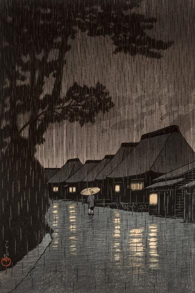 Evening rain falls on a rural street in Maekawa, in modern-day Kanagawa Prefecture. On our left, branches of tall shadowy trees reach out over the road; a row of traditional thatched-roof houses stands on our right, warm lights shining in their windows. The road running down the middle of the image is slick with rain, and steady rain continues to fall from a dim, overcast sky. A solitary pedestrian carries a bamboo umbrella over their head as they walk away from us.