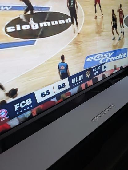 ALT-Text: Eine Fernsehszene eines Basketballspiels zwischen dem FC Bayern München (FCB) und ratiopharm Ulm (ULM). Der Spielstand zeigt 65:68 für Ulm bei 6:07 Minuten verbleibender Spielzeit im 4. Viertel. Auf dem Spielfeld sind Spieler und ein Schiedsrichter zu sehen. Die Aufnahme ist leicht schräg und zeigt den unteren Rand eines Fernsehers.